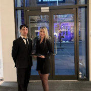 Two professionally dressed event staff members stand at the entrance of a modern venue, with one holding a tray and both smiling while welcoming guests