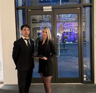 Two professionally dressed event staff members stand at the entrance of a modern venue, with one holding a tray and both smiling while welcoming guests
